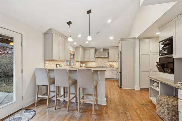 kitchen with pendant lighting, stainless steel fridge, light stone counters, and wall chimney range hood