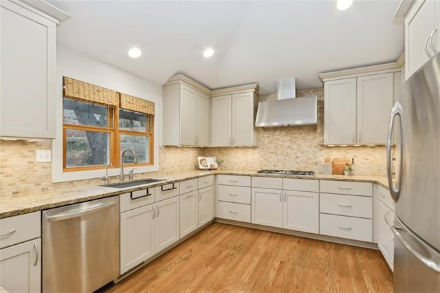 kitchen with sink, stainless steel appliances, light stone countertops, wall chimney range hood, and light hardwood / wood-style flooring