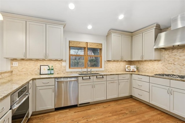 kitchen with sink, appliances with stainless steel finishes, light stone countertops, wall chimney exhaust hood, and light wood-type flooring