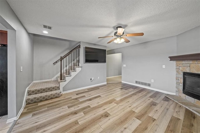 unfurnished living room with ceiling fan, a stone fireplace, a textured ceiling, and light hardwood / wood-style floors