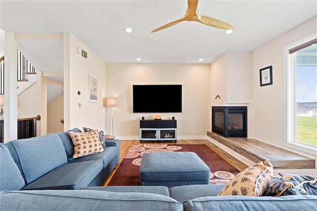living room with stairs, a glass covered fireplace, wood finished floors, recessed lighting, and a ceiling fan