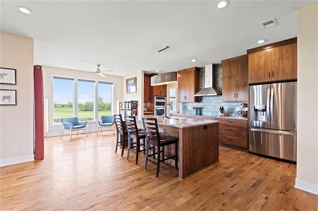 kitchen with a breakfast bar, wall chimney range hood, stainless steel refrigerator with ice dispenser, brown cabinets, and backsplash