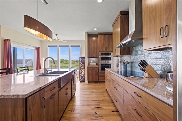 kitchen with a kitchen island with sink, a sink, double oven, black electric stovetop, and wall chimney exhaust hood