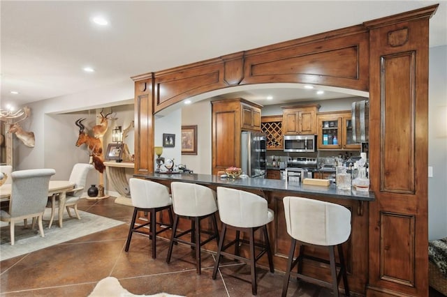 kitchen with dark countertops, appliances with stainless steel finishes, brown cabinets, a breakfast bar area, and a peninsula