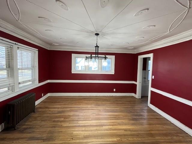 unfurnished dining area featuring a notable chandelier, wood finished floors, baseboards, ornamental molding, and radiator