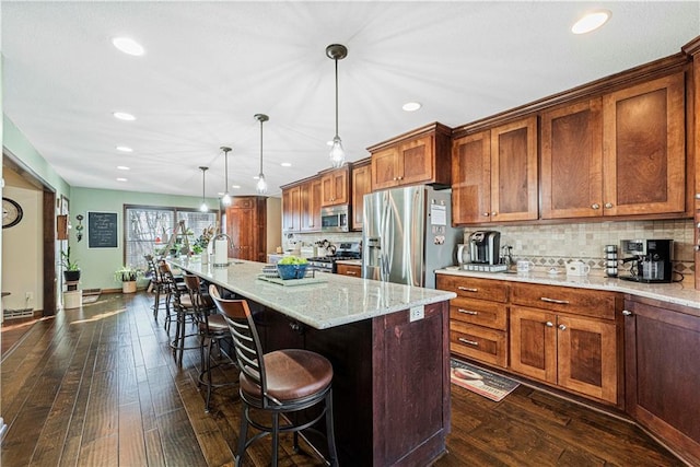 kitchen featuring pendant lighting, appliances with stainless steel finishes, a kitchen island with sink, light stone counters, and a kitchen bar