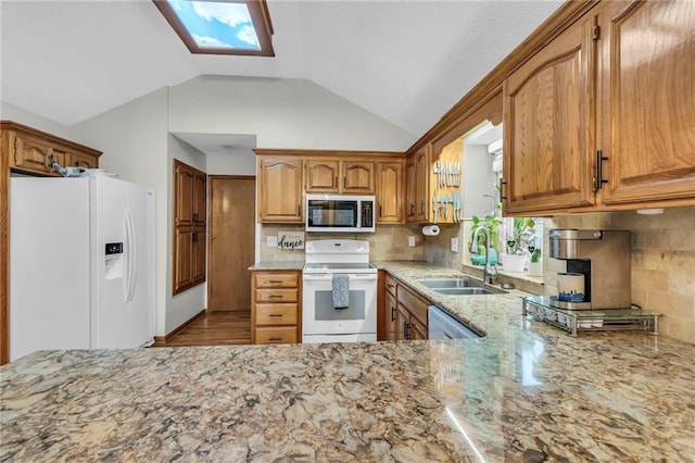 kitchen featuring vaulted ceiling with skylight, light stone counters, white appliances, a sink, and brown cabinets