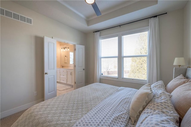 bedroom featuring a tray ceiling, light colored carpet, ensuite bathroom, baseboards, and visible vents