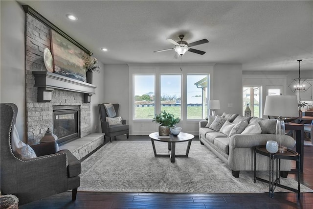 living room with wood-type flooring, a stone fireplace, ceiling fan with notable chandelier, and a textured ceiling