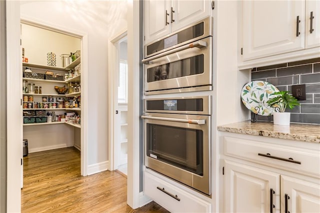 kitchen featuring stainless steel double oven, light hardwood / wood-style flooring, light stone counters, white cabinets, and backsplash