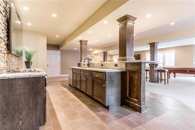 kitchen with a center island with sink, light stone countertops, stainless steel dishwasher, sink, and decorative columns