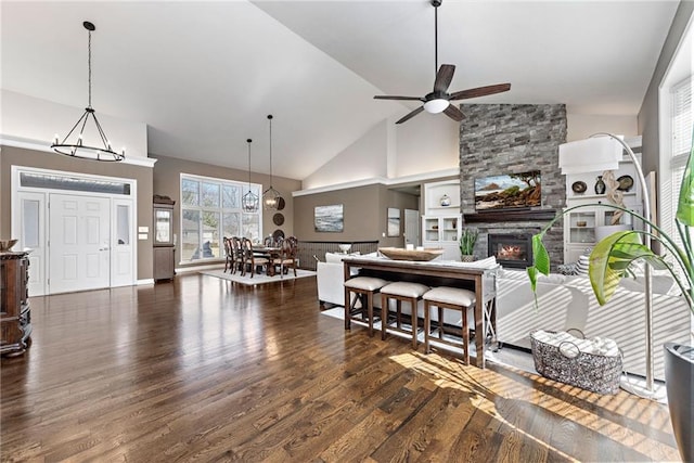 dining area featuring a stone fireplace, dark wood-type flooring, ceiling fan with notable chandelier, and high vaulted ceiling