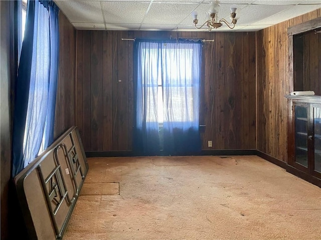 carpeted empty room with a drop ceiling, wooden walls, and a chandelier