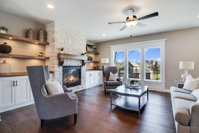 living room with a stone fireplace, dark wood-type flooring, and ceiling fan