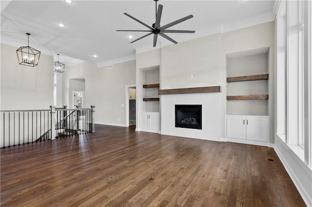 unfurnished living room featuring built in shelves, ornamental molding, ceiling fan with notable chandelier, and dark wood-type flooring