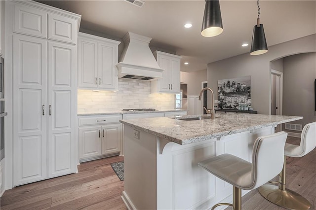 kitchen featuring white cabinetry, light stone countertops, an island with sink, and premium range hood