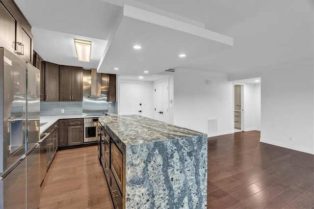 kitchen with wall chimney range hood, hardwood / wood-style flooring, stainless steel appliances, a center island, and light stone counters