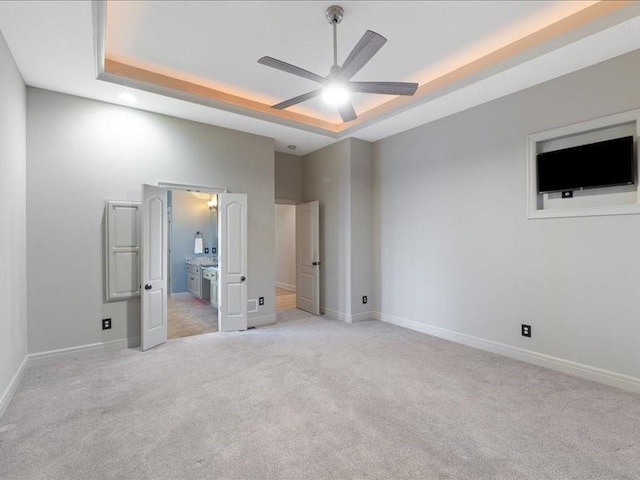 unfurnished bedroom with baseboards, a tray ceiling, ceiling fan, ensuite bathroom, and light colored carpet