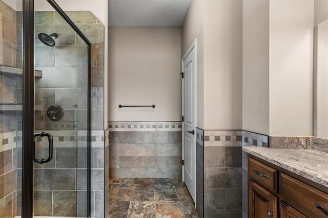 bathroom with a shower stall, vanity, wainscoting, stone finish floor, and tile walls