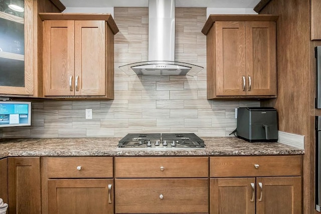 kitchen with light stone counters, stainless steel gas cooktop, decorative backsplash, brown cabinets, and wall chimney exhaust hood