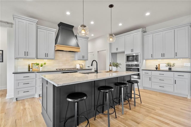 kitchen with decorative light fixtures, custom exhaust hood, stainless steel appliances, light stone countertops, and a center island with sink