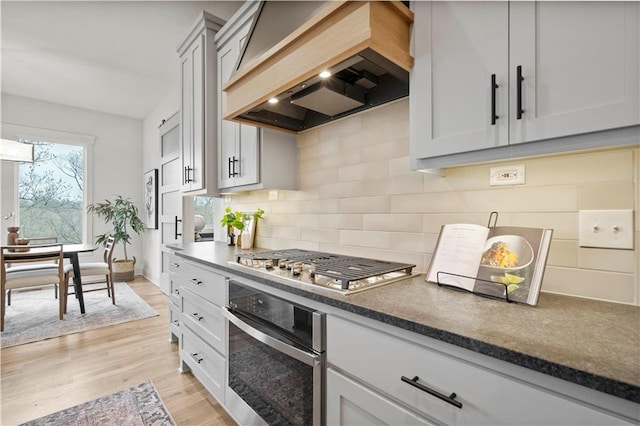 kitchen with stainless steel appliances, custom range hood, backsplash, and light wood-type flooring