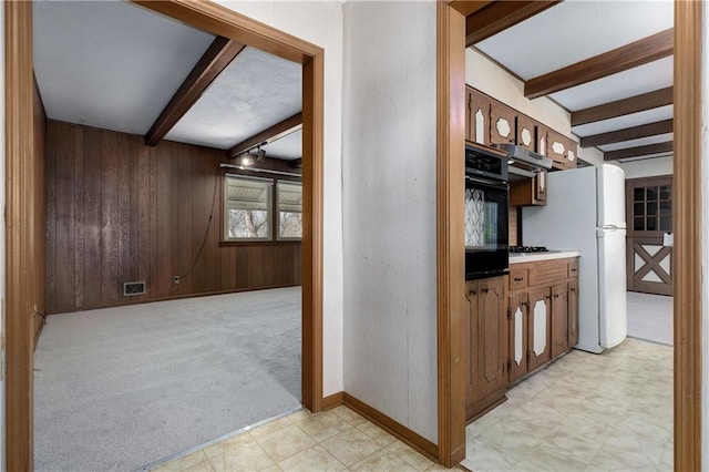 kitchen featuring black oven, beam ceiling, wooden walls, white refrigerator, and light colored carpet