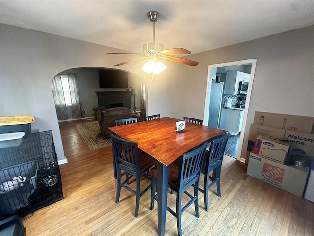 dining space featuring arched walkways, a fireplace, light wood finished floors, ceiling fan, and baseboards