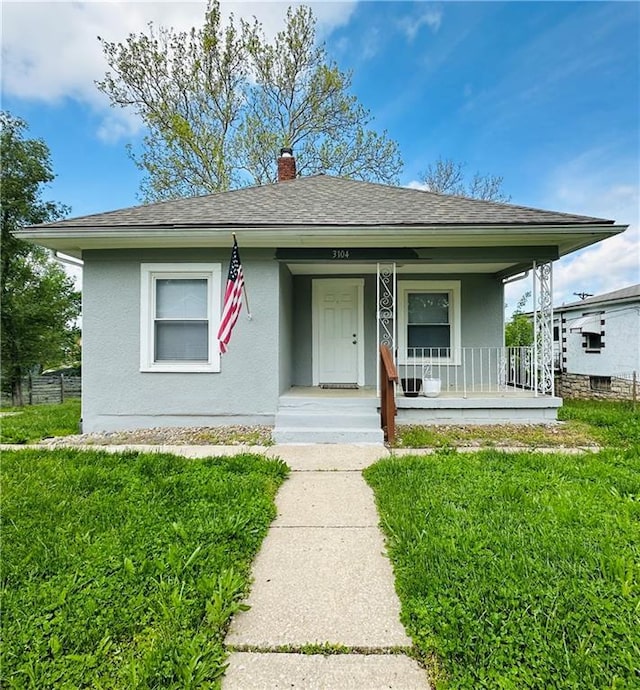 bungalow-style house with a shingled roof, stucco siding, a porch, a front yard, and a chimney