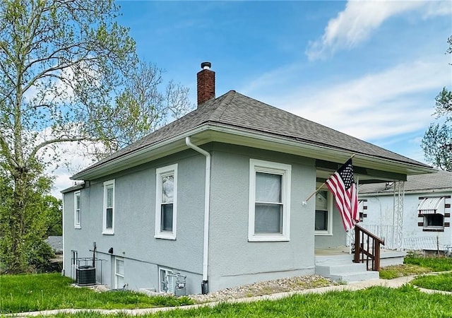 back of property with a shingled roof, stucco siding, central AC unit, a chimney, and a yard