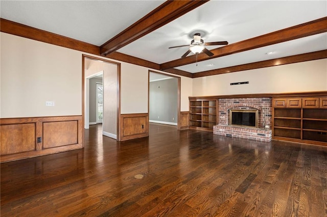 unfurnished living room featuring a ceiling fan, wood finished floors, beam ceiling, a fireplace, and wainscoting