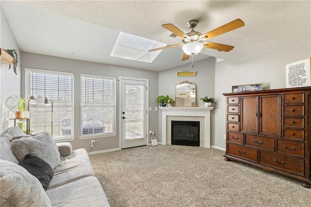 living area featuring a tiled fireplace, a ceiling fan, light carpet, vaulted ceiling with skylight, and baseboards