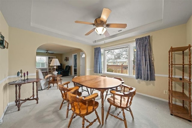 carpeted dining area featuring baseboards, visible vents, arched walkways, a ceiling fan, and a tray ceiling