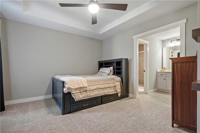 bedroom featuring a raised ceiling, light colored carpet, ensuite bathroom, a ceiling fan, and baseboards