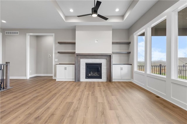 unfurnished living room featuring a fireplace, a raised ceiling, ceiling fan, and light wood-type flooring