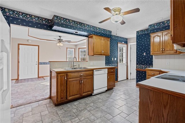 kitchen with brown cabinetry, wallpapered walls, white dishwasher, a sink, and light countertops