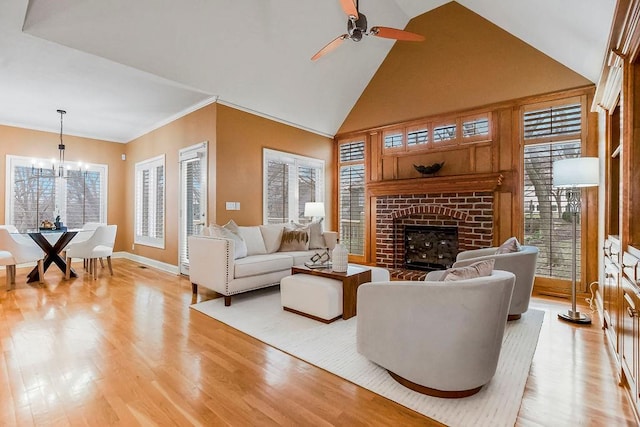 living room featuring high vaulted ceiling, ornamental molding, a brick fireplace, ceiling fan with notable chandelier, and light wood-type flooring