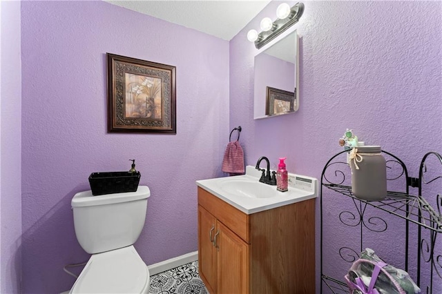 half bath featuring vanity, toilet, a textured wall, and baseboards