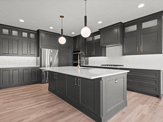 kitchen with a center island with sink, light countertops, hanging light fixtures, a sink, and light wood-type flooring