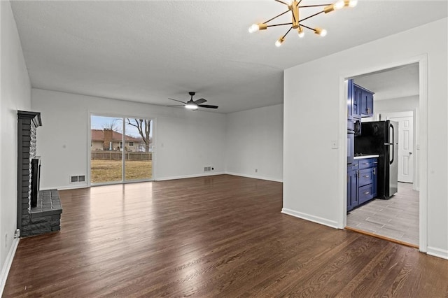 unfurnished living room with a brick fireplace, dark wood-type flooring, and ceiling fan with notable chandelier