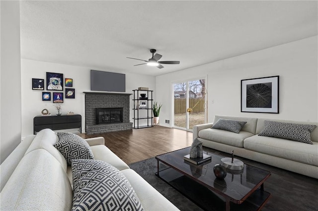 living room with a brick fireplace, dark wood-type flooring, and ceiling fan