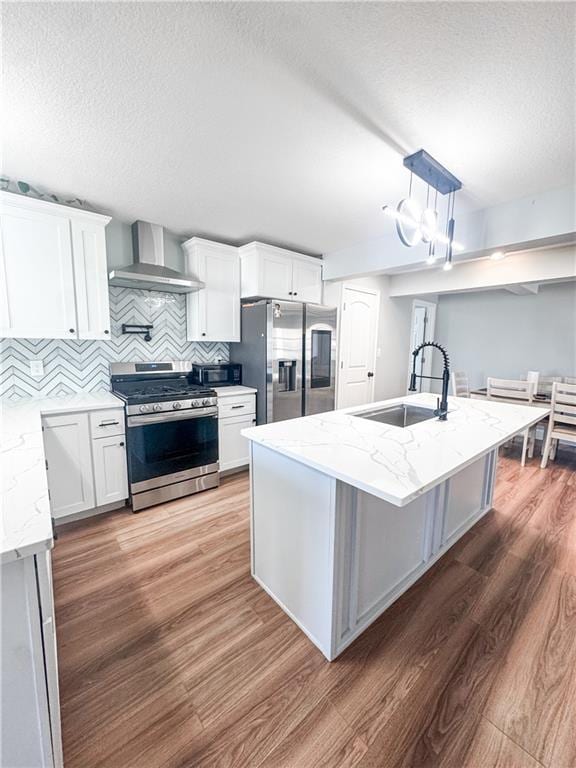 kitchen featuring wall chimney range hood, sink, stainless steel appliances, white cabinets, and a center island with sink