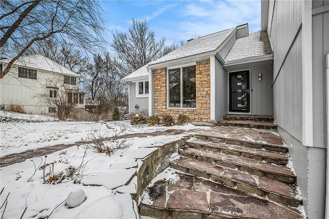 snow covered property entrance featuring a shingled roof and stone siding