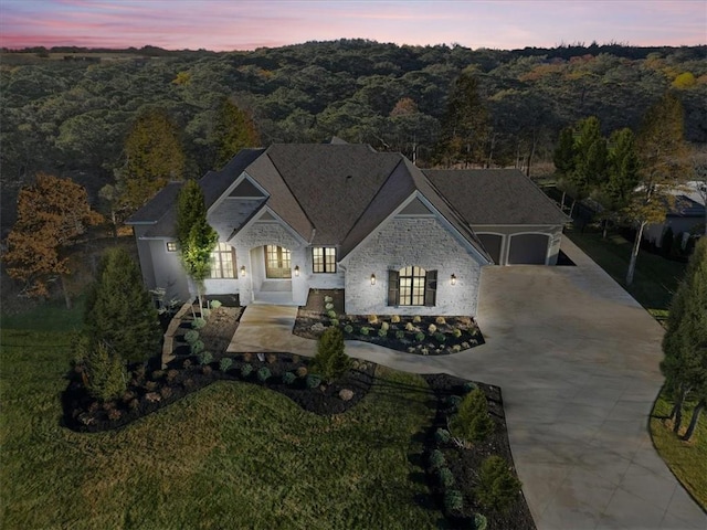 french country style house featuring a garage, a forest view, stone siding, and driveway