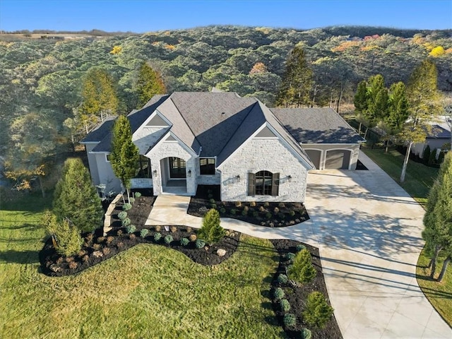 french provincial home with concrete driveway, a view of trees, a garage, stone siding, and a front lawn