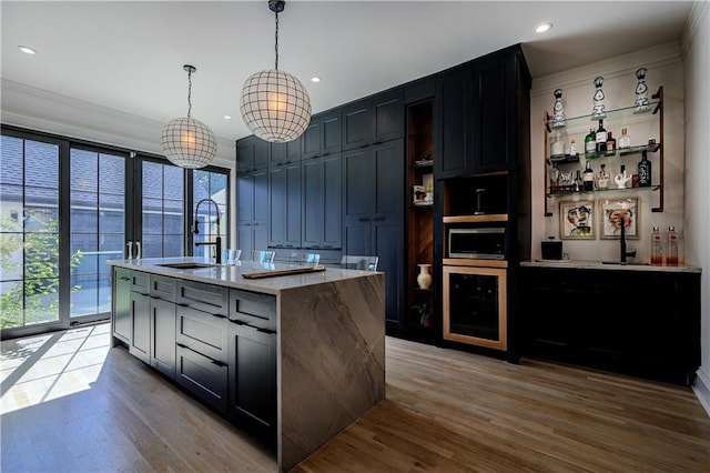 kitchen featuring beverage cooler, a center island with sink, a sink, decorative light fixtures, and wood finished floors