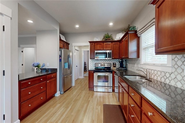 kitchen with stainless steel appliances, a sink, backsplash, light wood finished floors, and dark stone countertops