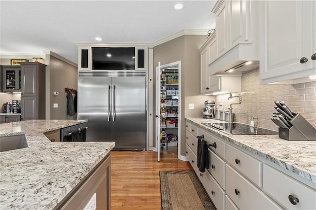 kitchen with light stone counters, black electric stovetop, built in refrigerator, white cabinetry, and glass insert cabinets