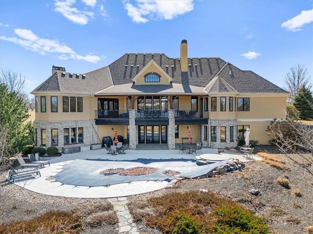 rear view of house with stucco siding, a chimney, a balcony, stone siding, and a patio