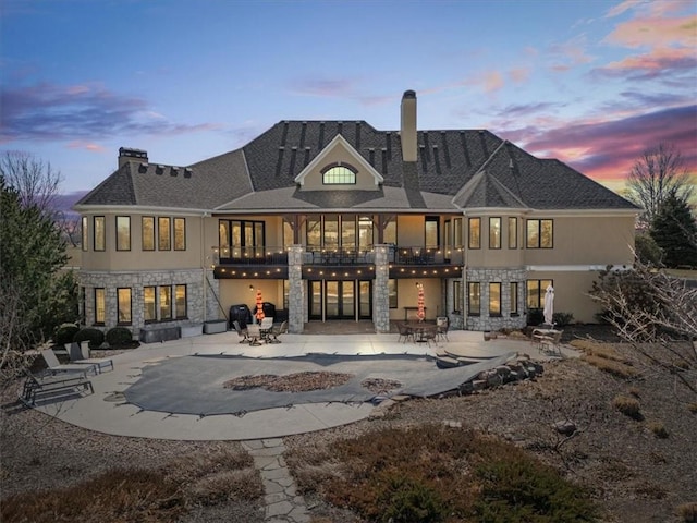 back of property at dusk with stone siding, a balcony, a patio, and a chimney